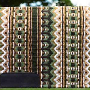 May include: A woven saddle pad with a geometric pattern in shades of green, brown, and white. The pad has a dark green border and a black leather patch at the bottom. The pad is displayed on a wooden fence.