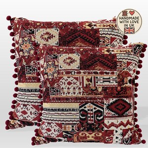 May include: Two decorative cushions with a patchwork design in shades of red, beige, and black. The cushions feature geometric and floral patterns, with a border of small red pom-poms. Handmade in the UK.