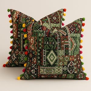 May include: Two square throw cushions with a patchwork design in green, red, and brown. The cushions feature a geometric pattern and are trimmed with colourful pom-poms. The cushions are designed for decorative purposes.