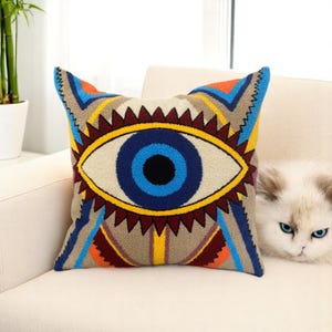 May include: Square decorative pillow featuring a large eye design with blue, yellow, and brown geometric patterns. The pillow has a cream-colored background and is placed on a white sofa. A white cat with blue eyes is visible in the background.