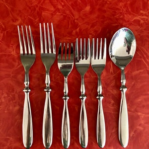 May include: A set of six silver-toned flatware pieces, including five forks and one spoon, arranged in a row on a red surface.