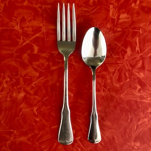 May include: A stainless steel fork and spoon set against a textured, reddish-orange background. The fork has four tines, and the spoon has a rounded bowl. The utensils are positioned side by side, creating a classic dining arrangement.