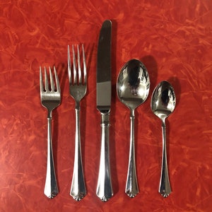 May include: A set of five silver-toned flatware pieces, including a knife, two forks, and two spoons, arranged in a row on a red textured surface.