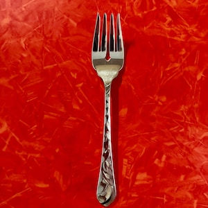 May include: A silver-toned metal fork featuring four tines and a decorative embossed leaf pattern along the length of the handle. The utensil has a polished, reflective finish and a tapered neck leading to the tines, presented against a bright red background.