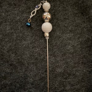 Puede incluir: Un palo de cuentas plateado y blanco con un amuleto de ojo turco azul. El palo mide aproximadamente 25 cm de largo y tiene una punta plateada.
