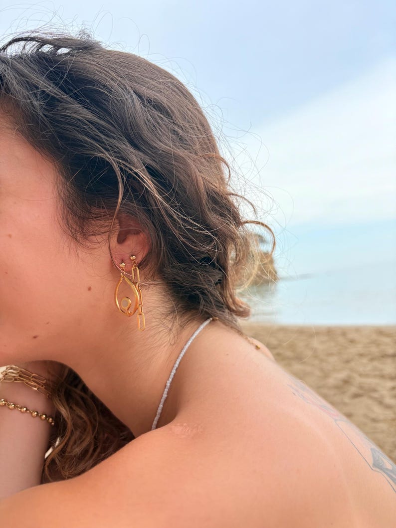 May include: A gold-toned earring set featuring a teardrop-shaped charm with a central white stone and a dangling rectangular chain link. The earring is secured with a small ball stud. Additionally, a gold-toned chain bracelet is visible on the wrist.