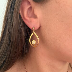May include: A gold-toned drop earring featuring an open teardrop-shaped frame with a small, circular white inlay centered at the base. The earring is attached to a simple gold ball stud post that secures it to the earlobe with a polished finish.