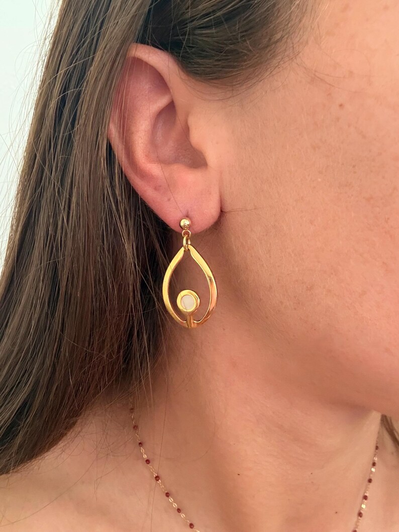 May include: A gold-toned drop earring featuring an open teardrop-shaped frame with a small, circular white inlay centered at the base. The earring is attached to a simple gold ball stud post that secures it to the earlobe with a polished finish.