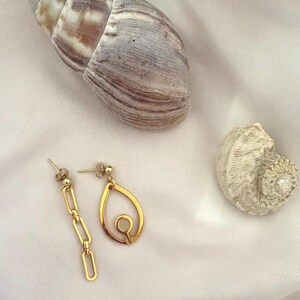 May include: A pair of mismatched gold-toned earrings. One earring features a vertical chain of three rectangular links, while the other earring consists of a teardrop-shaped frame with a small circular cutout in the center. Both have stud backings.