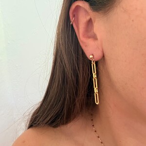 May include: A gold-toned earring featuring a small ball stud connected to a vertical drop composed of three elongated, rectangular chain links. A thin gold-toned hoop is also visible in the upper cartilage of the ear, along with a delicate necklace.