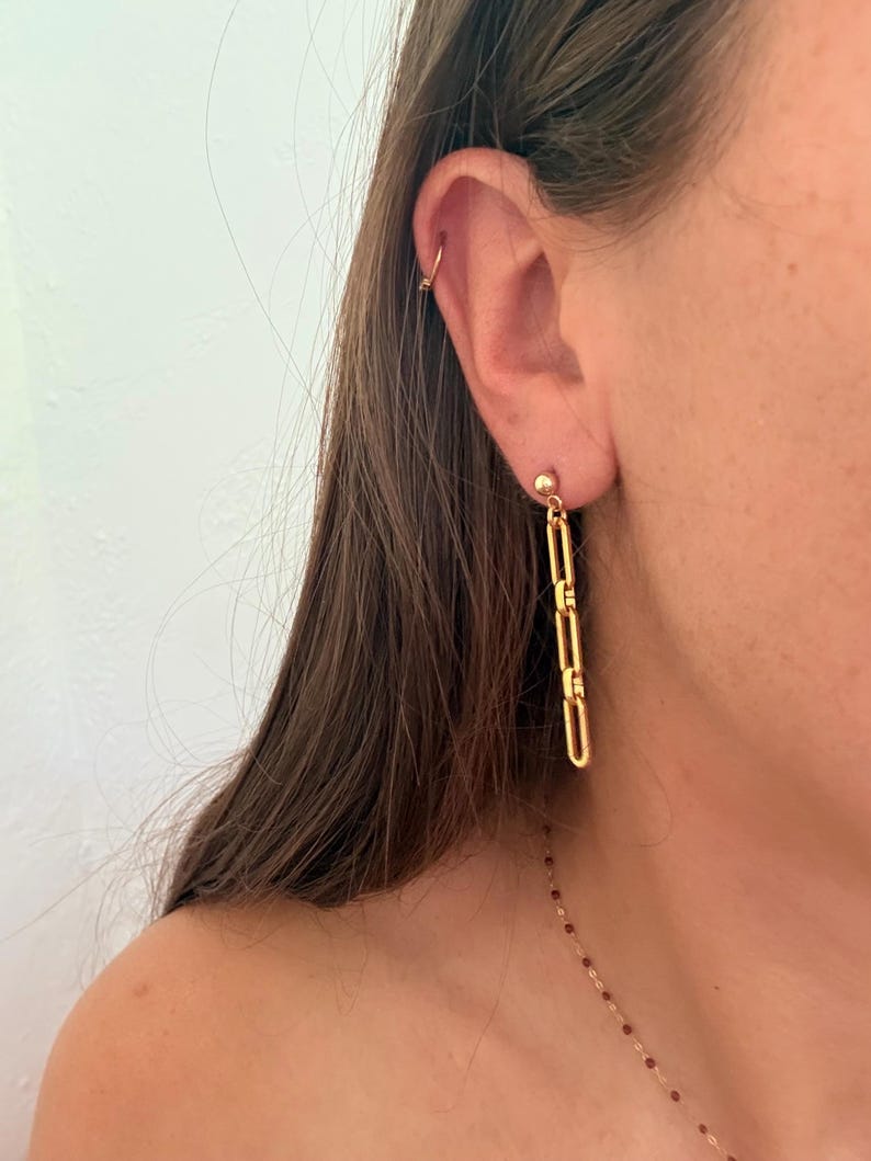 May include: A gold-toned earring featuring a small ball stud connected to a vertical drop composed of three elongated, rectangular chain links. A thin gold-toned hoop is also visible in the upper cartilage of the ear, along with a delicate necklace.