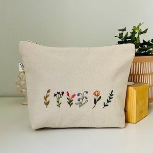 K&ouml;nnte beinhalten: Eine wei&szlig;e Canvas-Kosmetiktasche mit gestickten Blumen in Violett-, Rot- und Oranget&ouml;nen. Die Tasche liegt auf einer wei&szlig;en Oberfl&auml;che.