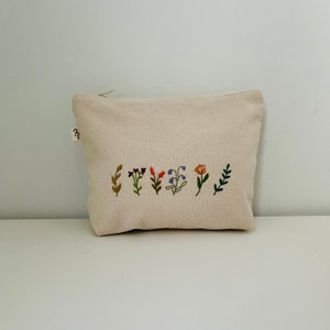 K&ouml;nnte beinhalten: Eine wei&szlig;e Canvas-Kosmetiktasche mit einer Reihe gestickter Blumen in verschiedenen Farben, darunter Rot, Orange, Lila und Gr&uuml;n.