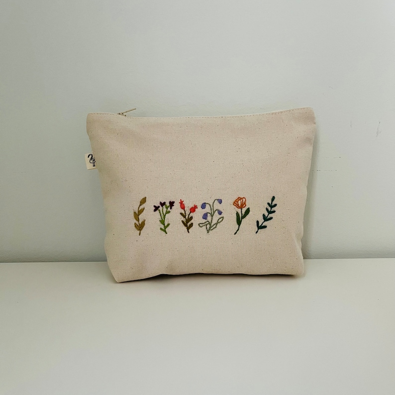 K&ouml;nnte beinhalten: Eine wei&szlig;e Canvas-Kosmetiktasche mit einer Reihe gestickter Blumen in verschiedenen Farben, darunter Rot, Orange, Lila und Gr&uuml;n.