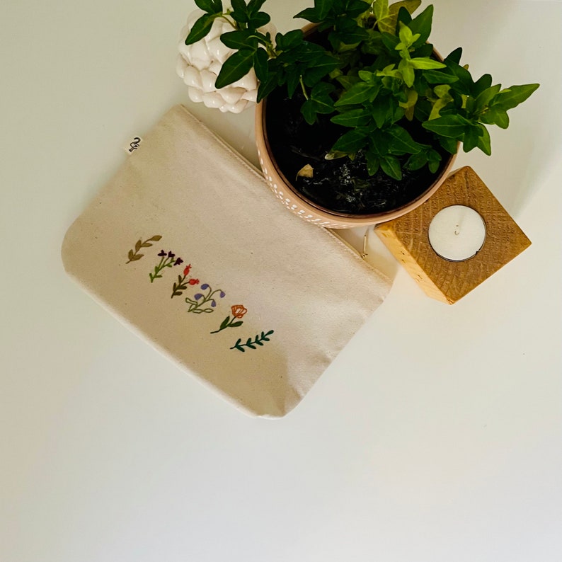 K&ouml;nnte beinhalten: Eine beige Stofftasche mit einem floralen Stickmuster. Die Tasche liegt auf einer wei&szlig;en Oberfl&auml;che.