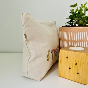 K&ouml;nnte beinhalten: Eine wei&szlig;e Canvas-Rei&szlig;verschlusstasche mit gestickten Blumen in Rosa-, Lila-, Gr&uuml;n- und Gelbt&ouml;nen. Die Tasche liegt auf einer wei&szlig;en Oberfl&auml;che neben einem Holzblock mit einer kleinen Kerze darauf und einem braunen Topf mit einer gr&uuml;nen Pflanze.