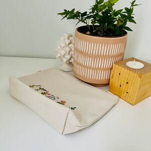 K&ouml;nnte beinhalten: Eine wei&szlig;e Stofftasche mit gestickten Blumen und dem Schriftzug "Good Vibes Only" in bunten F&auml;den. Die Tasche liegt auf einer wei&szlig;en Oberfl&auml;che neben einer Topfpflanze und einem Kerzenhalter aus Holz.