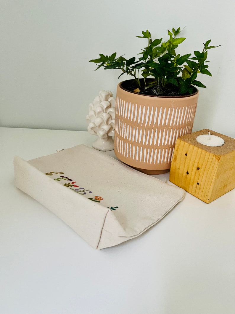 K&ouml;nnte beinhalten: Eine wei&szlig;e Stofftasche mit gestickten Blumen und dem Schriftzug "Good Vibes Only" in bunten F&auml;den. Die Tasche liegt auf einer wei&szlig;en Oberfl&auml;che neben einer Topfpflanze und einem Kerzenhalter aus Holz.