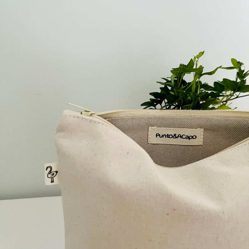 K&ouml;nnte beinhalten: Eine wei&szlig;e Canvas-Rei&szlig;verschlusstasche mit einem schwarzen Logo, das innen "Punto&A Capo" lautet. Die Tasche ist offen und ein kleines schwarzes Flamingo-Symbol ist au&szlig;en sichtbar.