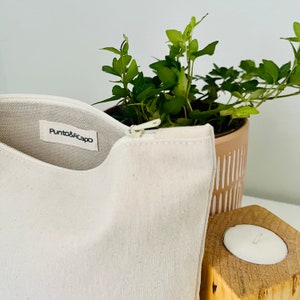 K&ouml;nnte beinhalten: Eine wei&szlig;e Canvas-Rei&szlig;verschlusstasche mit einem Etikett mit der Aufschrift "Punto&Acapo". Die Tasche liegt auf einer wei&szlig;en Oberfl&auml;che neben einer Topfpflanze und einem Kerzenhalter aus Holz.