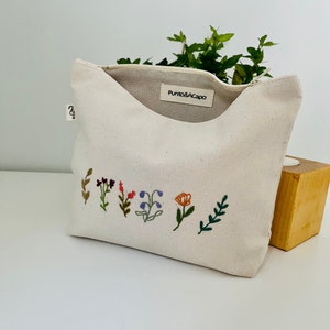 K&ouml;nnte beinhalten: Eine wei&szlig;e Canvas-Rei&szlig;verschlusstasche mit einem Etikett mit der Aufschrift "Punto&Acapo". Die Tasche hat ein florales Stickmuster mit bunten Blumen und Bl&auml;ttern.