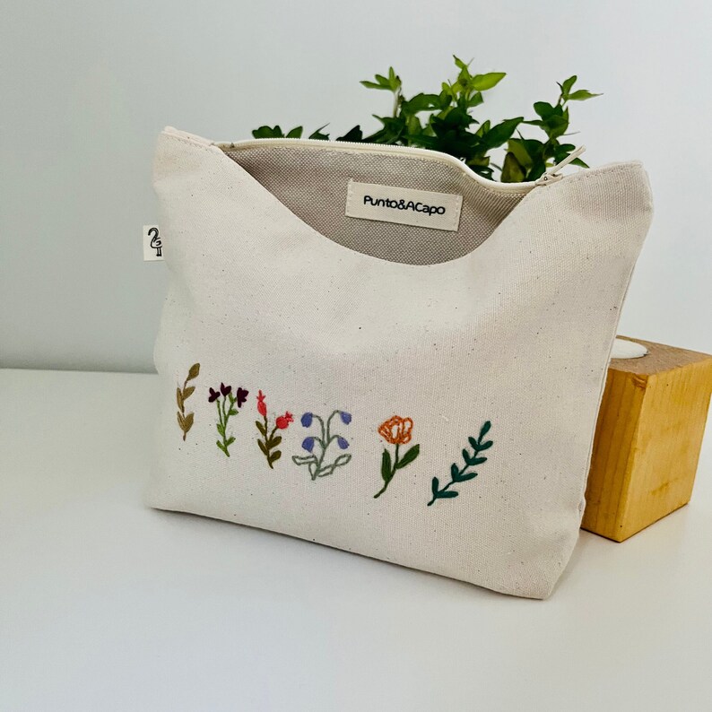 K&ouml;nnte beinhalten: Eine wei&szlig;e Canvas-Rei&szlig;verschlusstasche mit einem Etikett mit der Aufschrift "Punto&Acapo". Die Tasche hat ein florales Stickmuster mit bunten Blumen und Bl&auml;ttern.