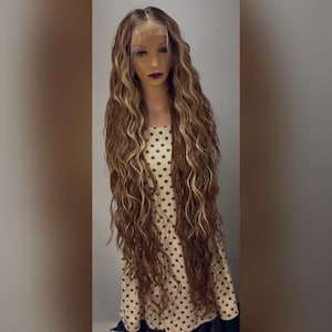 Puede incluir: Una larga peluca ondulada con mechas marrones y rubias. La peluca está peinada sobre una cabeza de maniquí con tez clara y lápiz labial oscuro. El cabello cae en cascada más allá de la cintura del maniquí, mostrando su longitud y textura. El maniquí lleva un vestido de lunares.