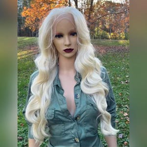 Peut inclure: Une perruque longue et ondulée, blonde platine, avec une frange en dentelle. La perruque est coiffée en vagues lâches et tombe sur les épaules. La perruque est portée par un mannequin avec une chemise verte.