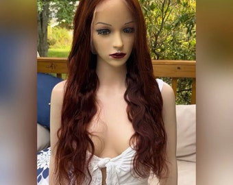 May include: A mannequin head with long, wavy auburn hair. The hair is styled with a deep part and falls to the shoulders. The mannequin is wearing a white top. The background is a natural outdoor setting with greenery.
