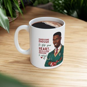May include: White ceramic mug with a black handle. The mug features a cartoon illustration of a Black man wearing a green doctor's coat and a red and white patterned scarf. The text on the mug reads "Handsome Enough to Stop Your Heart, Skilled Enough to Resist It".