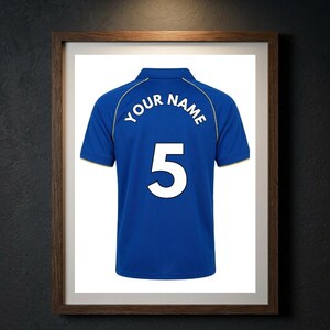 May include: A blue polo shirt featuring a white collar and sleeve trim, with the text "YOUR NAME" arched above the large white number "5" printed on the back. The shirt is displayed within a dark wooden frame against a dark textured wall.