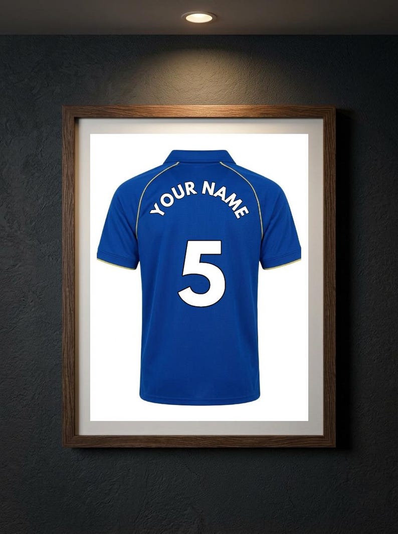 May include: A blue polo shirt featuring a white collar and sleeve trim, with the text "YOUR NAME" arched above the large white number "5" printed on the back. The shirt is displayed within a dark wooden frame against a dark textured wall.