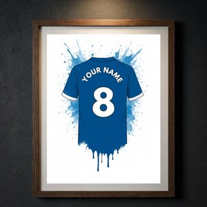 May include: A framed print featuring a blue sports jersey with white trim, the number 8, and the text YOUR NAME. The design includes a blue watercolor splash effect behind the jersey and a blue paint drip effect at the bottom hem of the shirt.