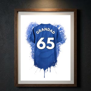 May include: A framed print featuring a blue sports jersey graphic with the word GRANDAD and the number 65 in white lettering. The design includes blue watercolor-style paint splatters and drips extending from the bottom of the jersey.