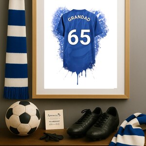 May include: A framed print featuring a blue sports jersey with the text GRANDAD and the number 65 in white. The jersey design includes a blue watercolor splash effect. The print is displayed alongside a soccer ball, black cleats, and blue and white scarves.