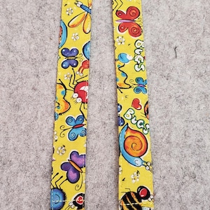 May include: Two yellow pacifier clips with a colorful bug pattern. The clips feature various insects like butterflies, ladybugs, and bees. Each clip has a metal clasp and a white loop.