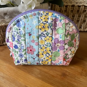 May include: A small, quilted cosmetic bag with a curved top and a purple zipper. The bag is made of fabric panels with floral patterns in various colors, including blue, yellow, pink, and purple. The bag is sitting on a wooden surface.