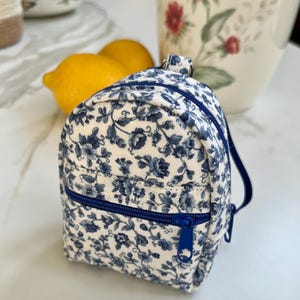 May include: A miniature backpack featuring a white fabric base with a blue floral pattern. The bag includes a front zippered pocket with a blue zipper pull, a main compartment with a blue zipper, and a small top loop for hanging or attachment.