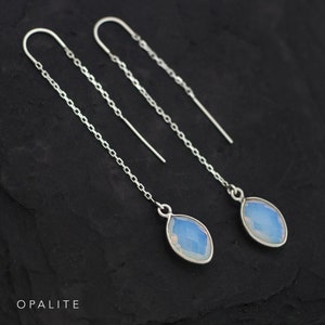 May include: A pair of silver threader earrings with opalescent teardrop-shaped stones. The earrings feature a delicate chain design and are displayed against a dark background. The word "OPALITE" is printed at the bottom of the image.