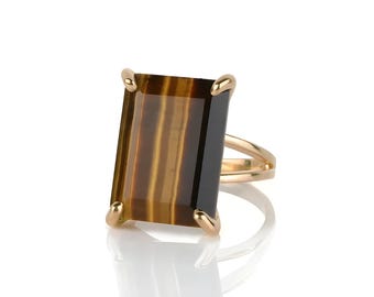 Op de afbeelding: Een gouden ring met een grote, rechthoekige tijgeroogsteen. De steen heeft bruine en gouden strepen. De ring heeft een split-schacht ontwerp en gouden klauwen die de steen vasthouden.