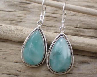 May include: A pair of silver teardrop earrings with a light blue gemstone center. The earrings have a decorative border and are suspended from a silver hook. The earrings are displayed on a wooden surface.