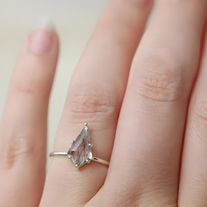 Peut inclure: Bague en argent ornée d'une pierre en forme de losange avec des inclusions internes. La pierre est translucide grise avec des lignes brun rougeâtre. La bague est montée sur un fin anneau en argent, présentant un design minimaliste.