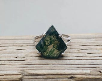 May include: A silver ring featuring a large, geometric moss agate stone. The stone is a deep green with intricate, natural patterns. The ring's band splits into two at the top, with small silver accents holding the stone in place. The ring is displayed on a wooden surface.