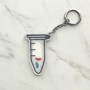 May include: Clear acrylic keychain shaped like a laboratory test tube, with a blue outline and a silver chain and keyring. The tube contains two DNA strands in red and blue.