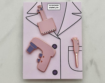 Può includere: Tre spille smaltate rosa a forma di strumenti da laboratorio, tra cui una pipetta, un dispositivo portatile e uno strumento più grande, sono disposte su una scheda rosa chiaro. La scheda presenta un contorno nero di un camice da laboratorio e il testo "microbe doodles enamel pins".