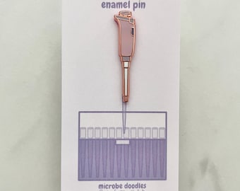 Può includere: Spilla in smalto rosa e rame con un design a pipetta, con la scritta "enamel pin" in alto. Sotto, un disegno di un rack di provette. Le parole "microbe doodles" sono in basso.