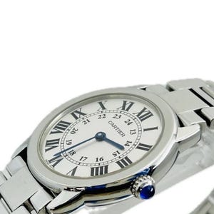 May include: A silver-toned Cartier watch with a white face and black Roman numeral hour markers. The watch features a blue second hand and a blue gemstone on the side. The watch band is also silver-toned.