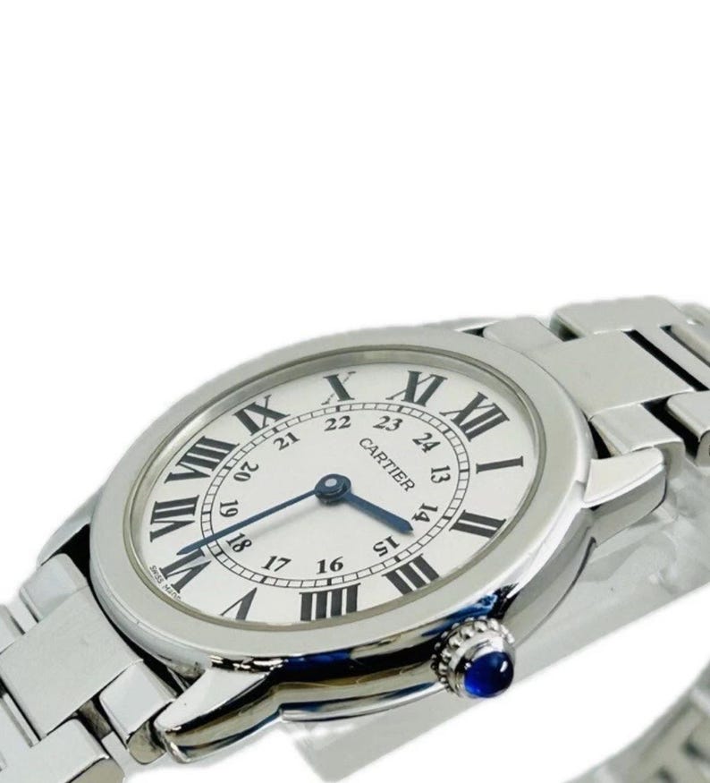 May include: A silver-toned Cartier watch with a white face and black Roman numeral hour markers. The watch features a blue second hand and a blue gemstone on the side. The watch band is also silver-toned.