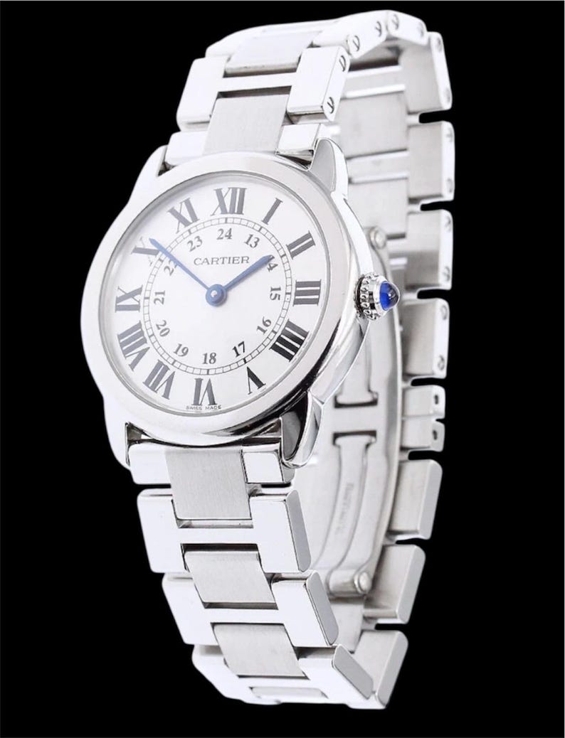 May include: A silver-toned wristwatch with a white face and black Roman numeral hour markers. The watch features a silver band and a blue gem on the side. The brand name "CARTIER" is printed on the watch face.