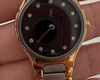 May include: A gold and silver two-tone wristwatch with a black face and diamond hour markers. The watch face also features the Yves Saint Laurent logo. The watch band is a two-tone metal link band. "Made in France" is printed on the watch face.